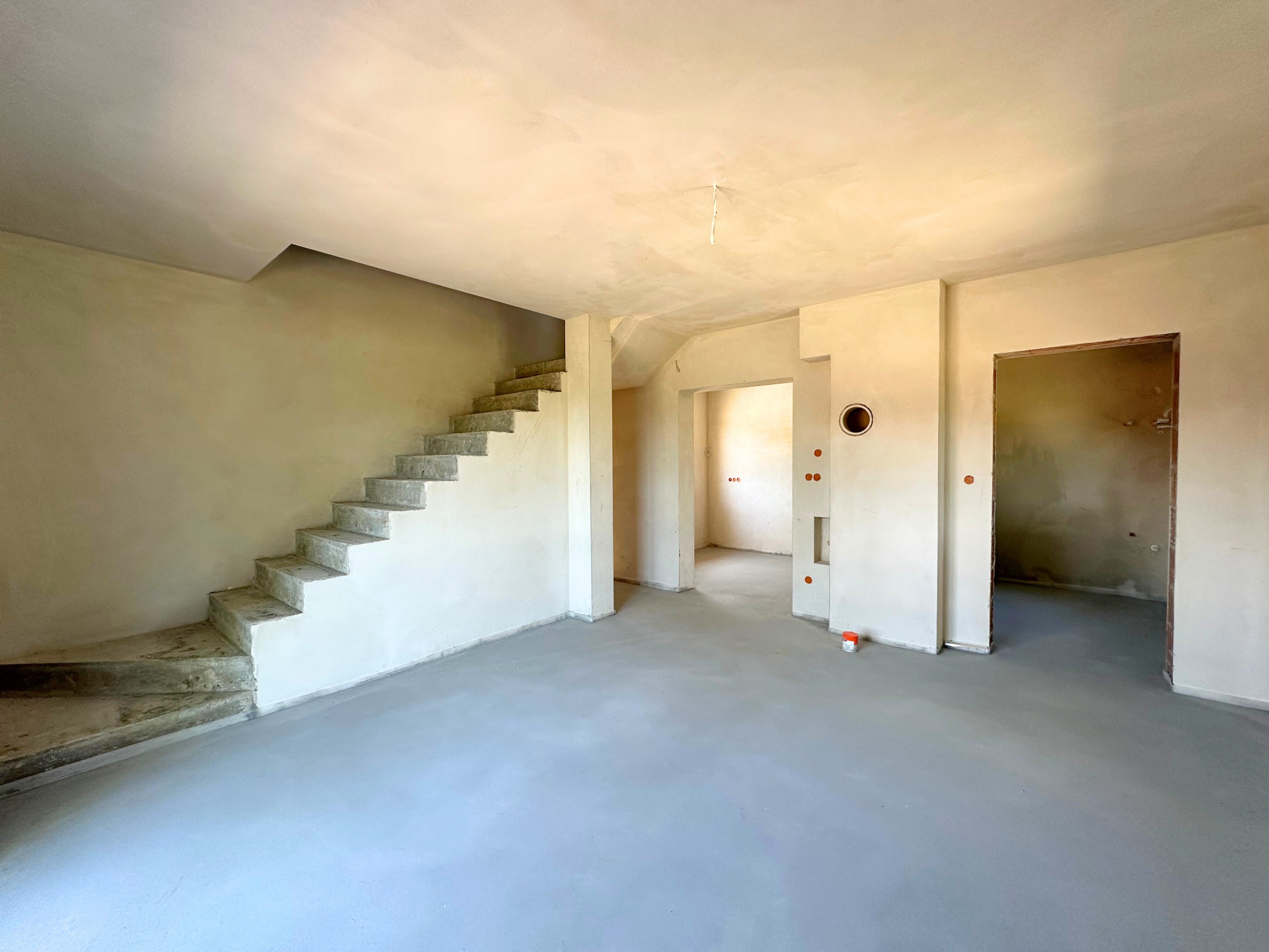 Surowe wnętrze domu w trakcie wykańczania: schody betonowe, wylewka na podłodze, ściany w tynku, widoczne otwory drzwiowe i przejścia. Minimalistyczny, surowy styl.