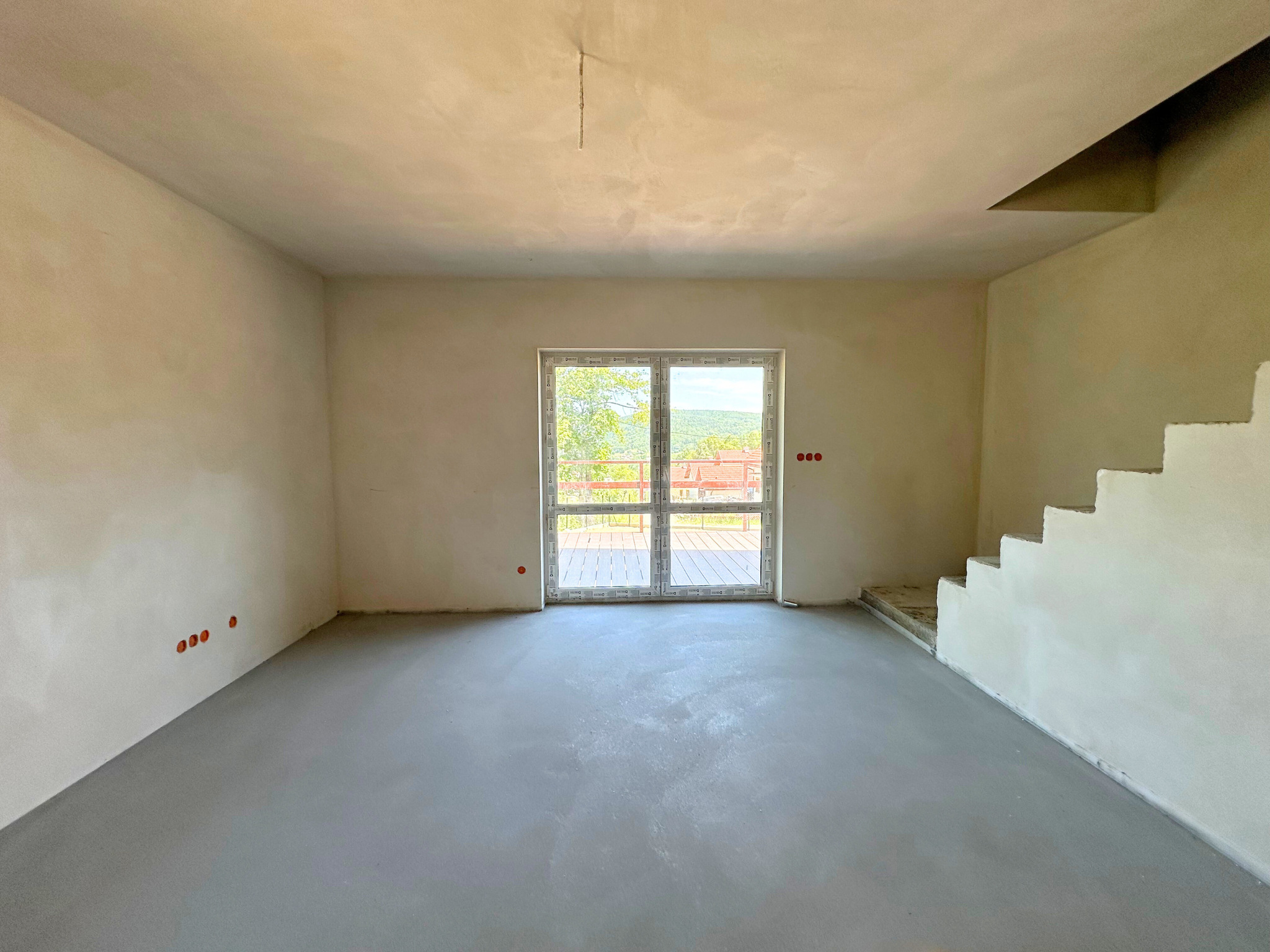 Surowe wnętrze z wylaną posadzką, przygotowane do dalszych prac wykończeniowych. Widoczne ściany po tynkowaniu, okno balkonowe i surowe schody.