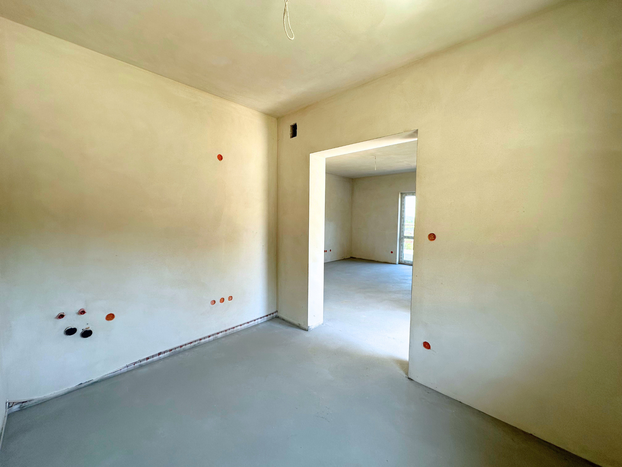 Wnętrze w stanie deweloperskim: gładkie ściany, wylewka, przygotowane punkty elektryczne, widok na kolejne pomieszczenie z oknem. Minimalizm i surowość wykończenia.