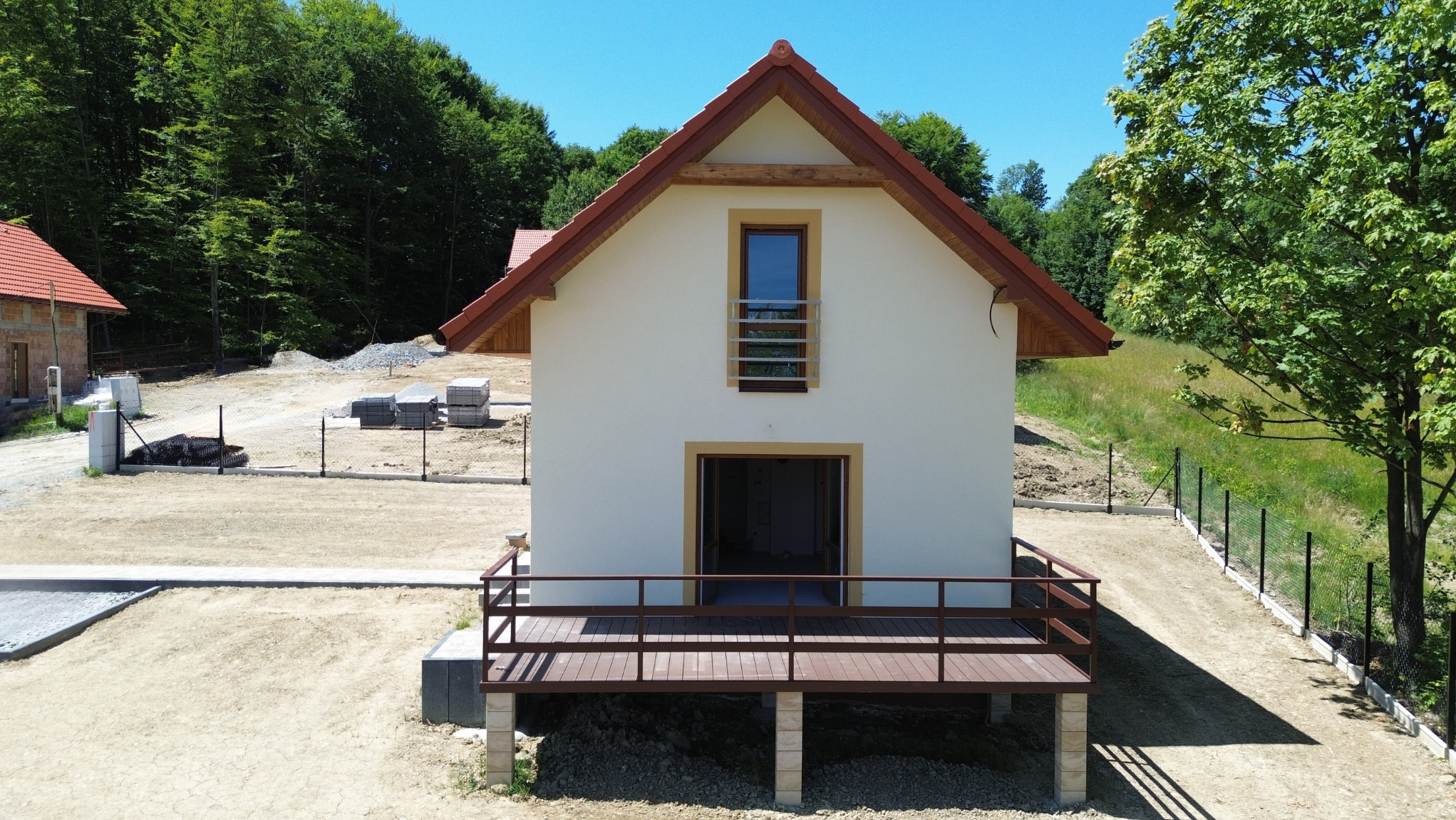 Nowoczesny dom jednorodzinny w stanie deweloperskim z drewnianym tarasem, ogrodzeniem i wyplantowaną działką, otoczony zielenią lasu i błękitnym niebem.