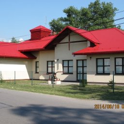 Dolomatex - Budynek z czerwonym dachem z blachy trapezowej, beżową elewacją i ciemnymi oknami, otoczony zielonym trawnikiem i ogrodzeniem z siatki. Widok z poziomu ulicy.