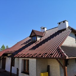 Dolomatex - Dom jednorodzinny z brązowym dachem z blachy trapezowej, białymi kominami i oknem dachowym typu lukarna. Widoczne rynny i fragment elewacji w jasnym kolorze.