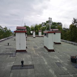 Dolomatex - Płaski dach pokryty papą z kilkoma kominami w kolorze białym z brązowymi akcentami. W tle drzewa i pochmurne niebo. Widoczne narzędzia dekarskie.