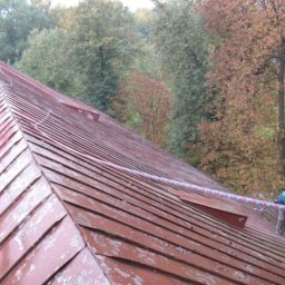 Dolomatex - Czerwony dach z blachy trapezowej, czyszczony myjką ciśnieniową przez pracownika w niebieskiej kurtce, zabezpieczonego liną. W tle drzewa.