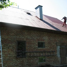 Dolomatex - Pracownik w uprzęży na blaszanym dachu czyści go myjką ciśnieniową. Obok ceglana ściana budynku z oknami i kominem. Stoi drabina.