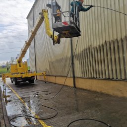 Dolomatex - Pracownik na podnośniku koszowym czyści elewację budynku przemysłowego myjką ciśnieniową, widoczne efekty czyszczenia. Żółty podnośnik MAN na mokrym placu.