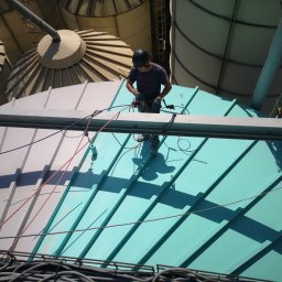 Dolomatex - Pracownik w kasku, zabezpieczony linami, stoi na pomalowanym na turkusowo dachu z blachy trapezowej. W tle silosy i kable. Ujęcie z góry.