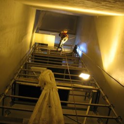 Dolomatex - Pracownik na rusztowaniu maluje wysokie wnętrze klatki schodowej. Widok z dołu, oświetlenie halogenowe, ściany w jasnym odcieniu. Praca na wysokościach.
