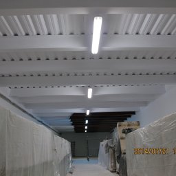 Dolomatex - Białe, pomalowane belki stropowe i blaszany dach w hali magazynowej, oświetlone lampami jarzeniowymi. Część towaru zabezpieczona folią. Widok z dołu.