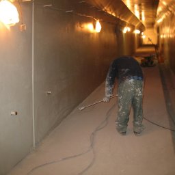 Dolomatex - Pracownik w kombinezonie maluje wnętrze tunelu natryskowo, widoczne oświetlenie i sprzęt malarski w tle. Świeżo pomalowane ściany i podłoga.