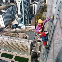 Dolomatex - Pracownik w kasku, na wysokości, myje okna wieżowca w Warszawie. Widok z góry, z perspektywy stopy pracownika. W tle panorama miasta.