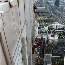 Dolomatex - Pracownicy wysokościowi w kombinezonach, zabezpieczeni linami, malują elewację wieżowca w centrum miasta. Widok z góry na panoramę miejską.