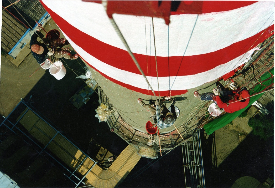 Alpiniści malują wieżę w biało-czerwone pasy, widok z góry. Prace na wysokości, liny, wiadra z farbą. Nietypowe ujęcie z perspektywy pracujących.