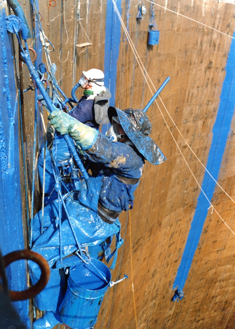 Dwóch pracowników w kombinezonach maluje na niebiesko wysoką, pionową powierzchnię. Używają lin i zabezpieczeń. Widoczne wiadro z farbą i narzędzia.