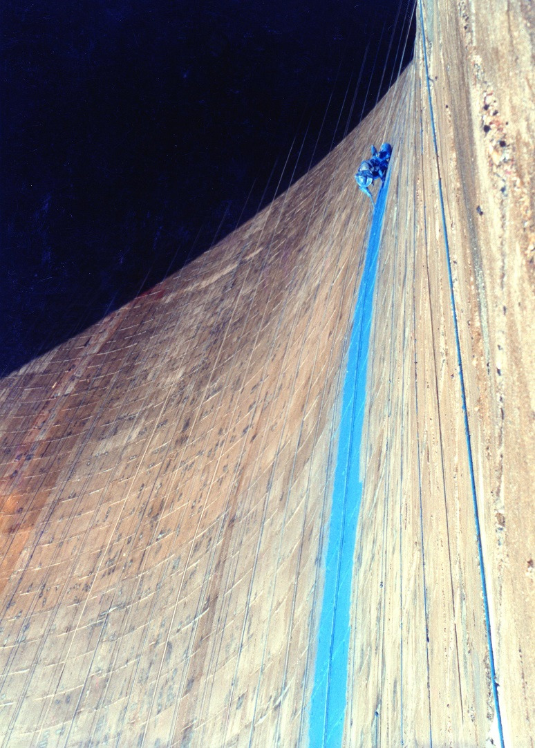 Pracownik w uprzęży maluje pionowy pas na zakrzywionej, zewnętrznej ścianie budynku. Widoczne liny i faktura ściany. Ujęcie z dołu, podkreślające skalę.