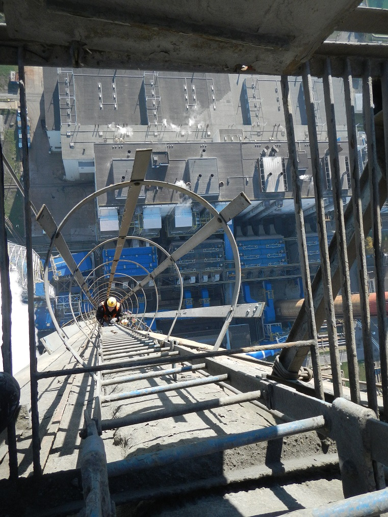 Pracownik w kasku na wysokiej, metalowej konstrukcji z drabinką, z widokiem na budynki przemysłowe. Ujęcie z góry, podkreślające wysokość i trudność dostępu.