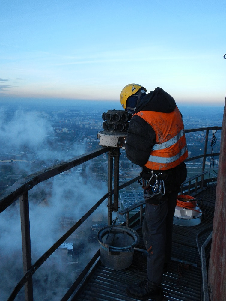 Pracownik w kasku i kamizelce na wysokości, prawdopodobnie na wieży, wykonuje prace konserwacyjne. Widoczny sprzęt i panorama miasta w tle.