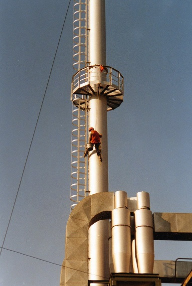 Pracownik w czerwonej kurtce maluje wysoki, srebrny komin przemysłowy z drabinką i platformą, zabezpieczony linami. Widoczne elementy instalacji przemysłowej.
