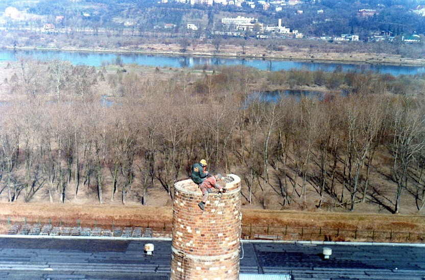 Dwóch robotników na szczycie ceglanego komina, prawdopodobnie podczas naprawy. W tle widok na rzekę i zabudowania miejskie. Ujęcie z góry, z dachu budynku.