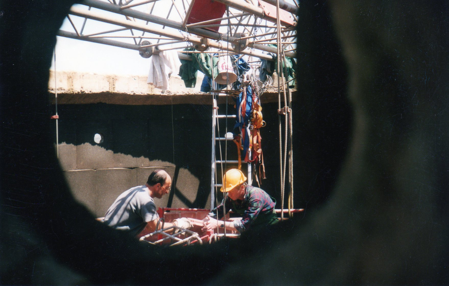 Dwóch robotników na rusztowaniu, jeden w kasku, pracuje przy czarnej ścianie z otworem, widok przez okrągły otwór.