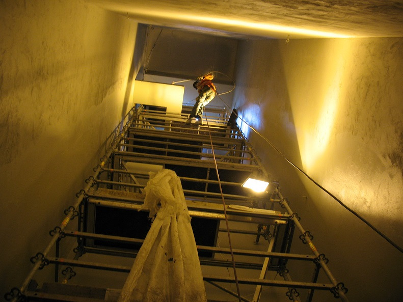 Pracownik na rusztowaniu maluje wysokie wnętrze klatki schodowej. Widok z dołu, oświetlenie halogenowe, ściany w jasnym odcieniu. Praca na wysokościach.