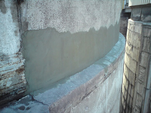 Fragment naprawionej, betonowej elewacji budynku z widocznymi śladami wcześniejszych uszkodzeń i świeżo nałożoną warstwą zaprawy wyrównującej.