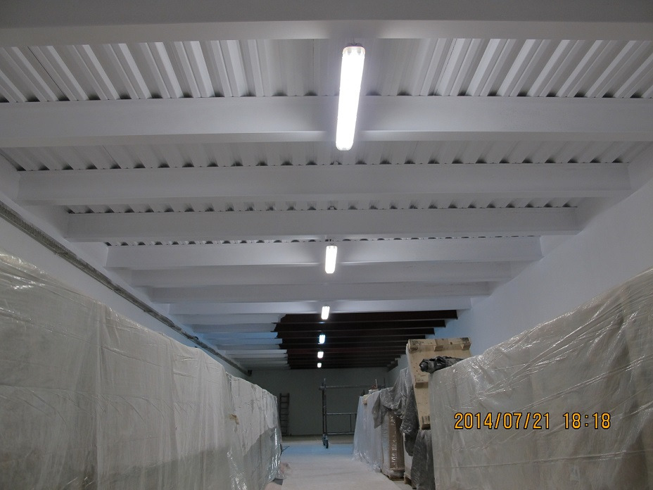 Białe, pomalowane belki stropowe i blaszany dach w hali magazynowej, oświetlone lampami jarzeniowymi. Część towaru zabezpieczona folią. Widok z dołu.