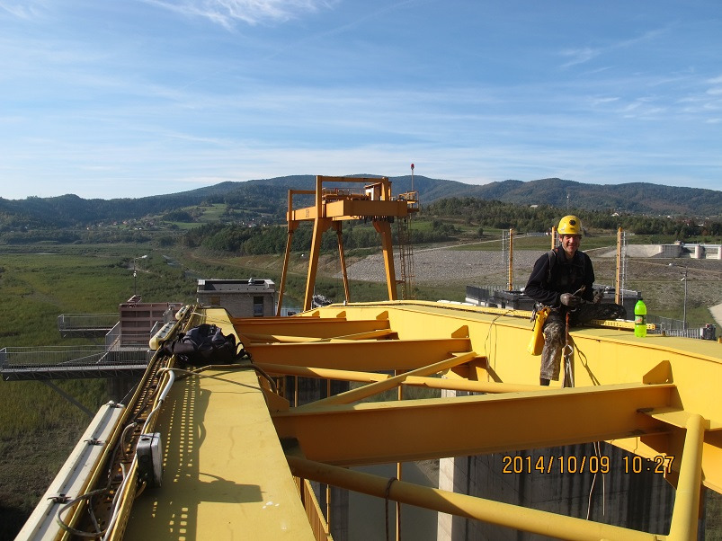 Pracownik w kasku na żółtej konstrukcji stalowej, prawdopodobnie mostowej, z widokiem na góry i zbiornik wodny w tle. Data 2014/10/09 na pierwszym planie.