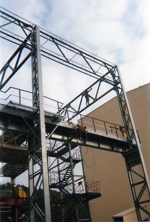 Prace na wysokościach: Dwóch robotników zabezpiecza metalową konstrukcję, jeden na górze, drugi na platformie, z widocznymi schodami i elementami konstrukcyjnymi.