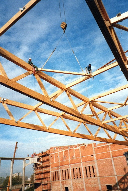Konstrukcja drewniana dachu w trakcie montażu, robotnicy na belkach, w tle budynek z cegły i dźwig. Błękitne niebo w tle.