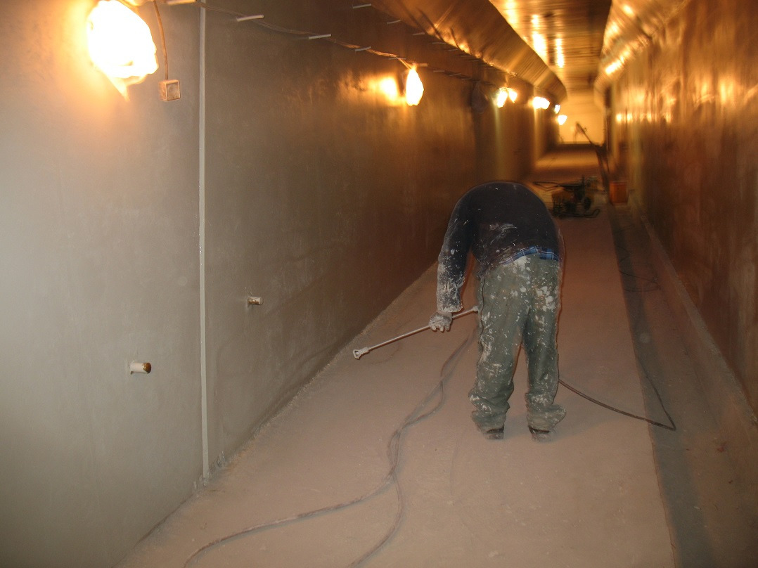 Pracownik w kombinezonie maluje wnętrze tunelu natryskowo, widoczne oświetlenie i sprzęt malarski w tle. Świeżo pomalowane ściany i podłoga.