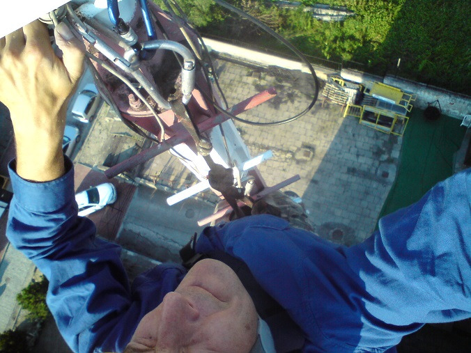 Pracownik w niebieskim kombinezonie, z perspektywy z góry, podczas pracy na wysokości na dachu budynku. Widoczne narzędzia i fragment dachu z zielonym elementem.