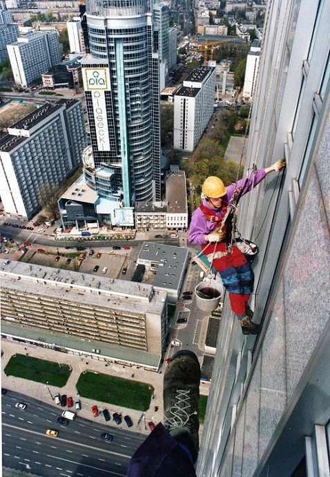 Pracownik w kasku, na wysokości, myje okna wieżowca w Warszawie. Widok z góry, z perspektywy stopy pracownika. W tle panorama miasta.