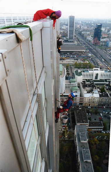 Pracownicy wysokościowi w kombinezonach, zabezpieczeni linami, malują elewację wieżowca w centrum miasta. Widok z góry na panoramę miejską.
