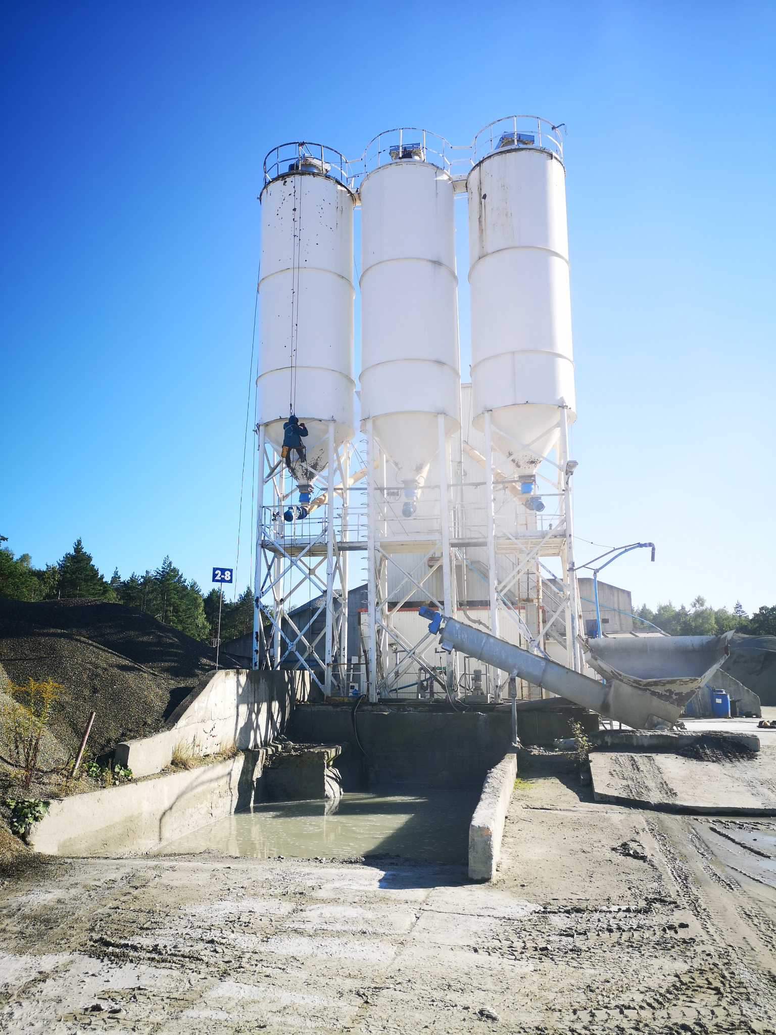 Białe silosy cementowe na tle błękitnego nieba, z pracownikiem na wysokościach i śladami opon na ziemi. Widoczny element infrastruktury przemysłowej.