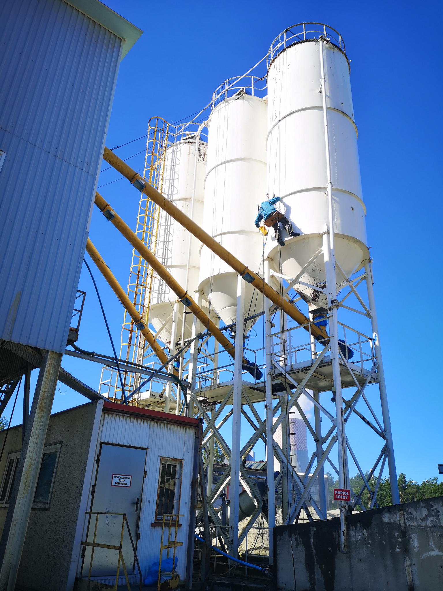Pracownik w uprzęży, na wysokości, czyści silosy na tle błękitnego nieba. Widoczne żółte rury i konstrukcja wsporcza. Tabliczka ostrzegawcza w dolnej części kadru.