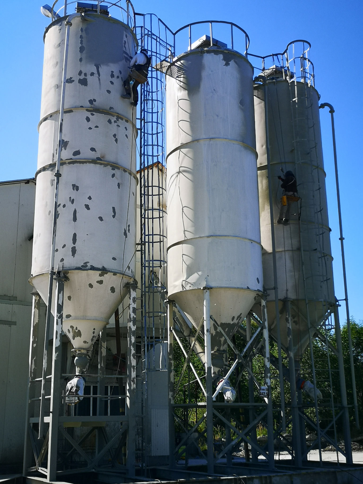 Pracownicy na linach malują silosy przemysłowe na tle błękitnego nieba. Widoczne ślady starej farby i drabiny. Praca na wysokościach, zabezpieczenia.