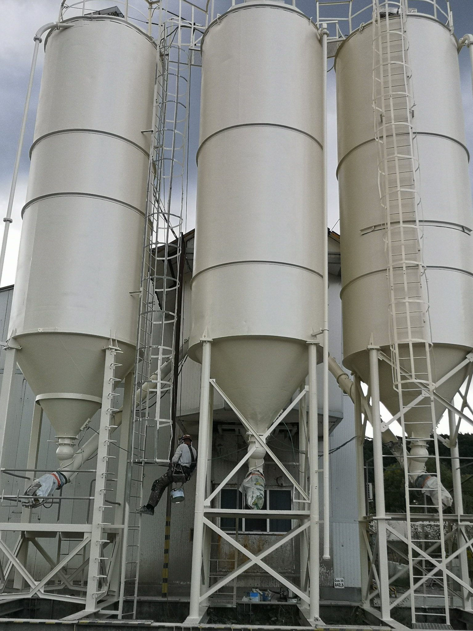 Pracownik na drabinie maluje silosy przemysłowe na biało. Widoczne trzy silosy z drabinkami i konstrukcją wsporczą. Praca na wysokości, zabezpieczenia.