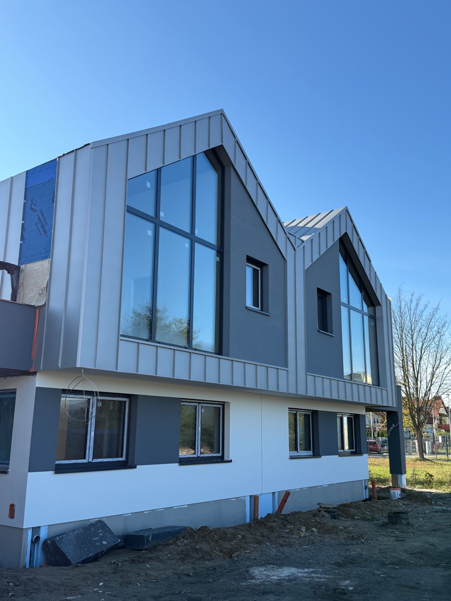 Nowoczesny dom w trakcie prac elewacyjnych: szara blacha na dachu i ścianach, duże okna, widoczne materiały izolacyjne. Częściowo otynkowana elewacja.