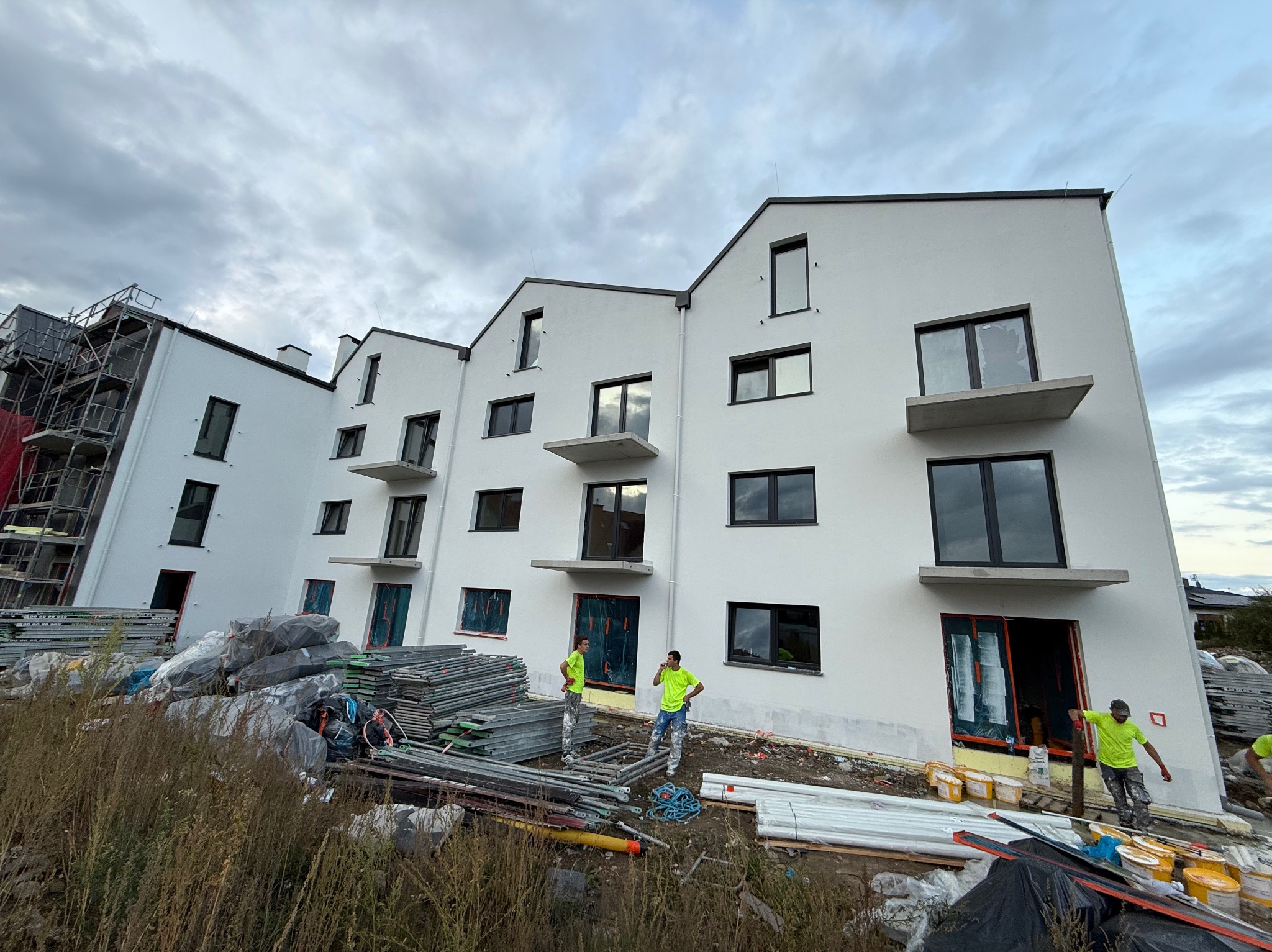 Nowoczesny budynek wielorodzinny z białą elewacją w trakcie prac wykończeniowych. Widoczne rusztowania, materiały budowlane i pracownicy w jaskrawych koszulkach.