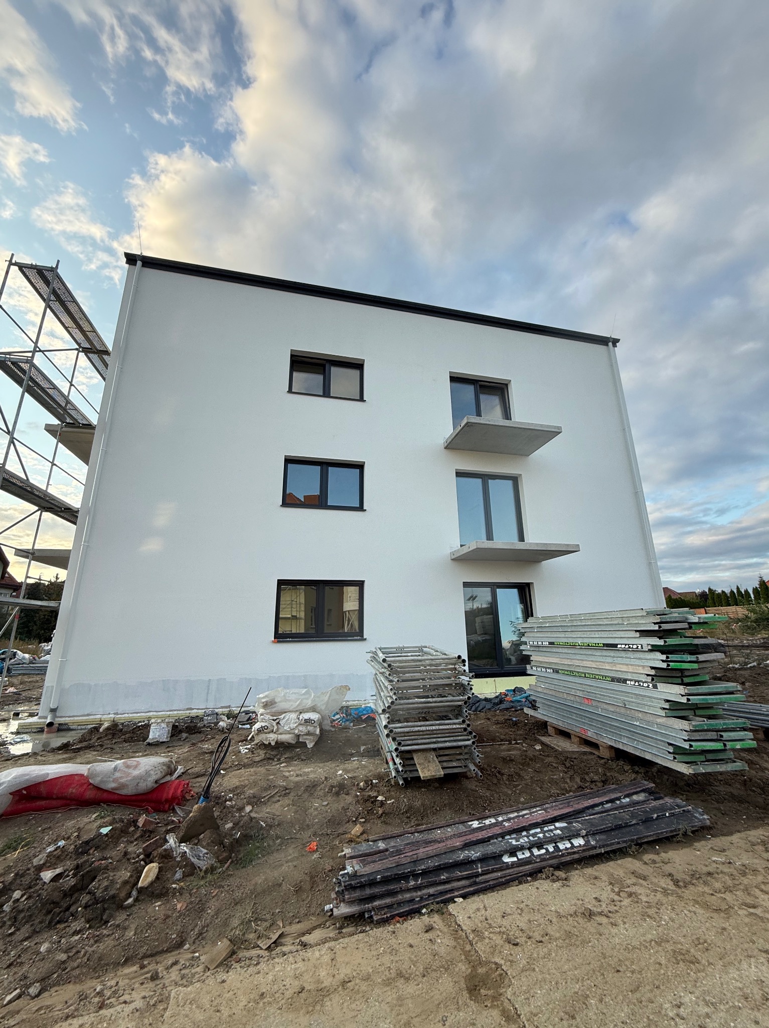 Nowoczesny, trzypiętrowy budynek z białą elewacją i balkonami, w trakcie prac wykończeniowych. Widoczne rusztowania i materiały budowlane. Ujęcie z dołu, pochmurne niebo.