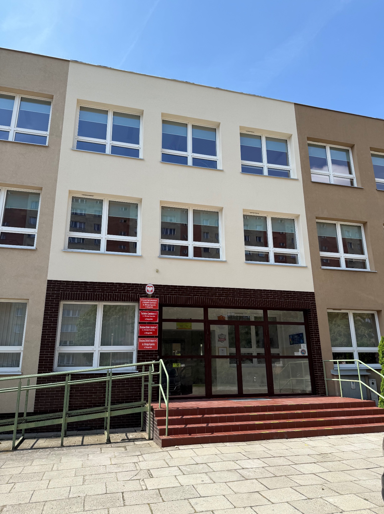 Budynek szkoły po renowacji elewacji, z widocznymi dwoma odcieniami beżu i brązu. Wejście z rampą dla niepełnosprawnych i tabliczkami informacyjnymi.