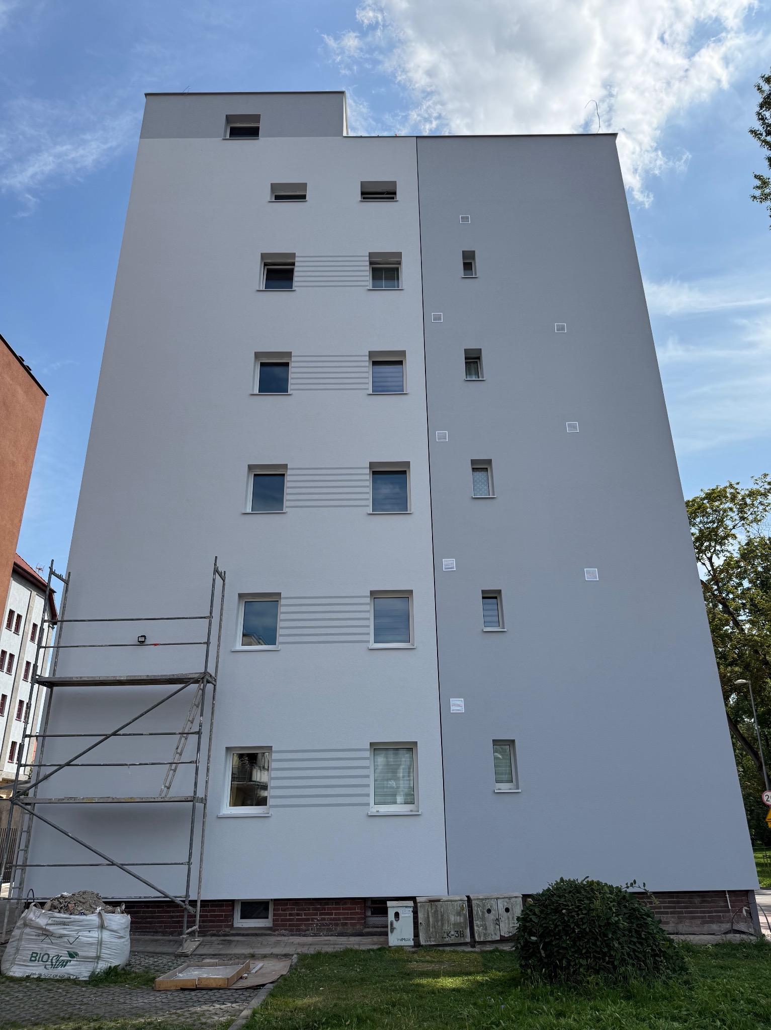 Wysoki budynek po renowacji elewacji w odcieniach szarości, z widocznymi rusztowaniami i workiem z materiałami budowlanymi u dołu. Czyste, błękitne niebo w tle.