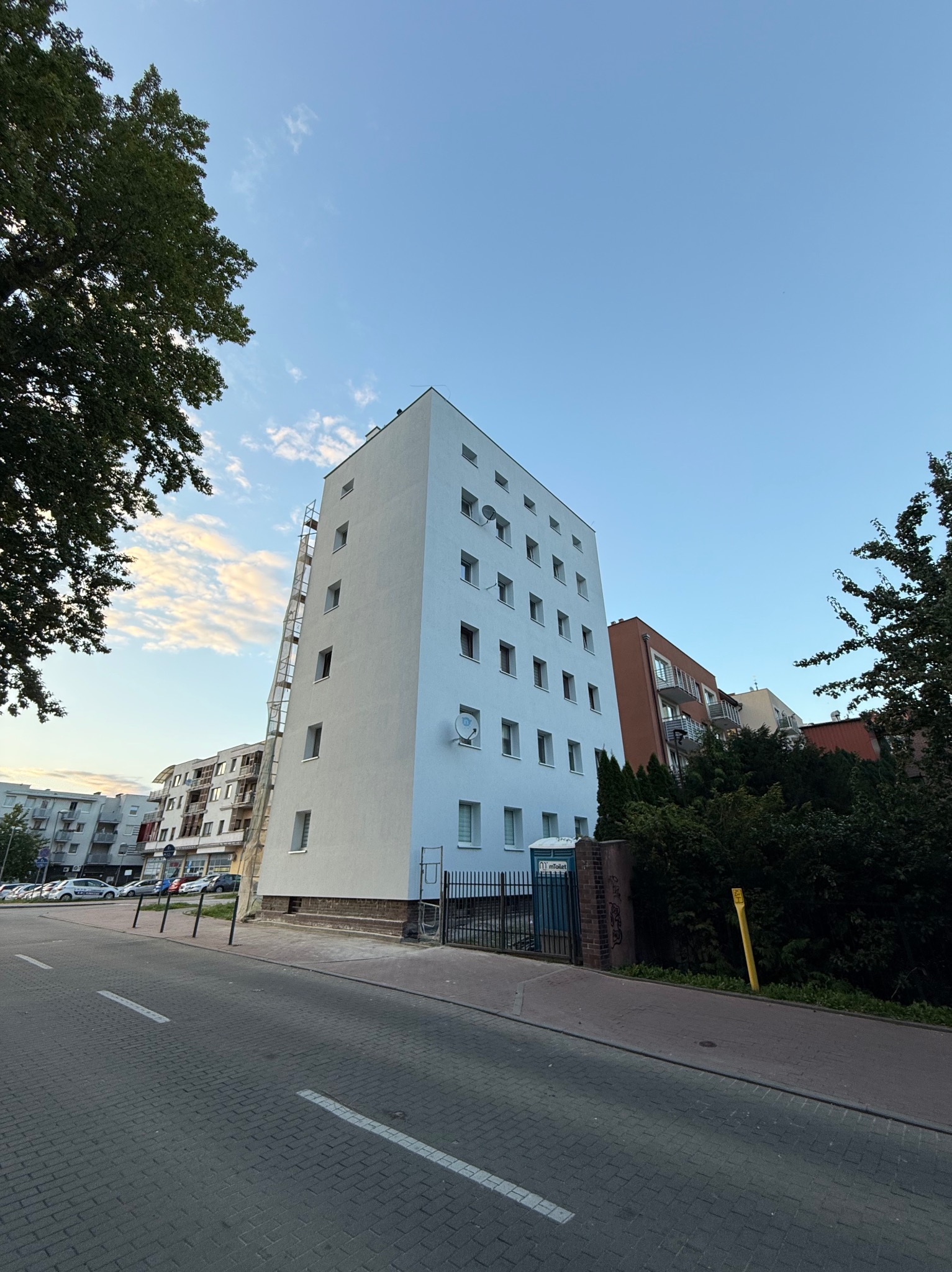 Wysoki budynek po renowacji elewacji, z podnośnikiem koszowym przy ścianie. Widok z dołu, błękitne niebo w tle. Biała, odświeżona fasada.