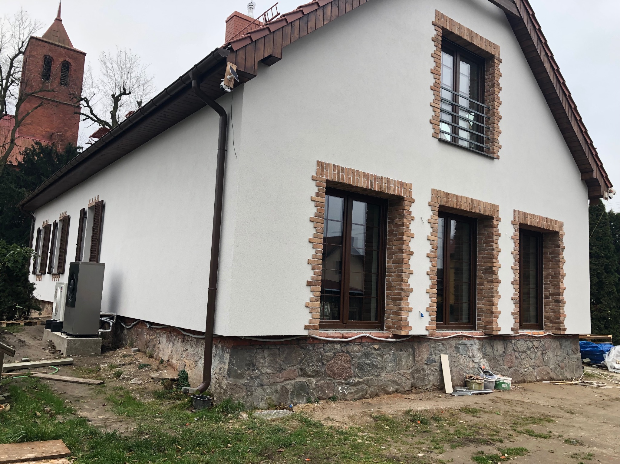 Dom z białą elewacją i ceglanymi obramowaniami okien, widoczny z boku, z pompą ciepła na zewnątrz. W tle ceglana wieża kościoła.