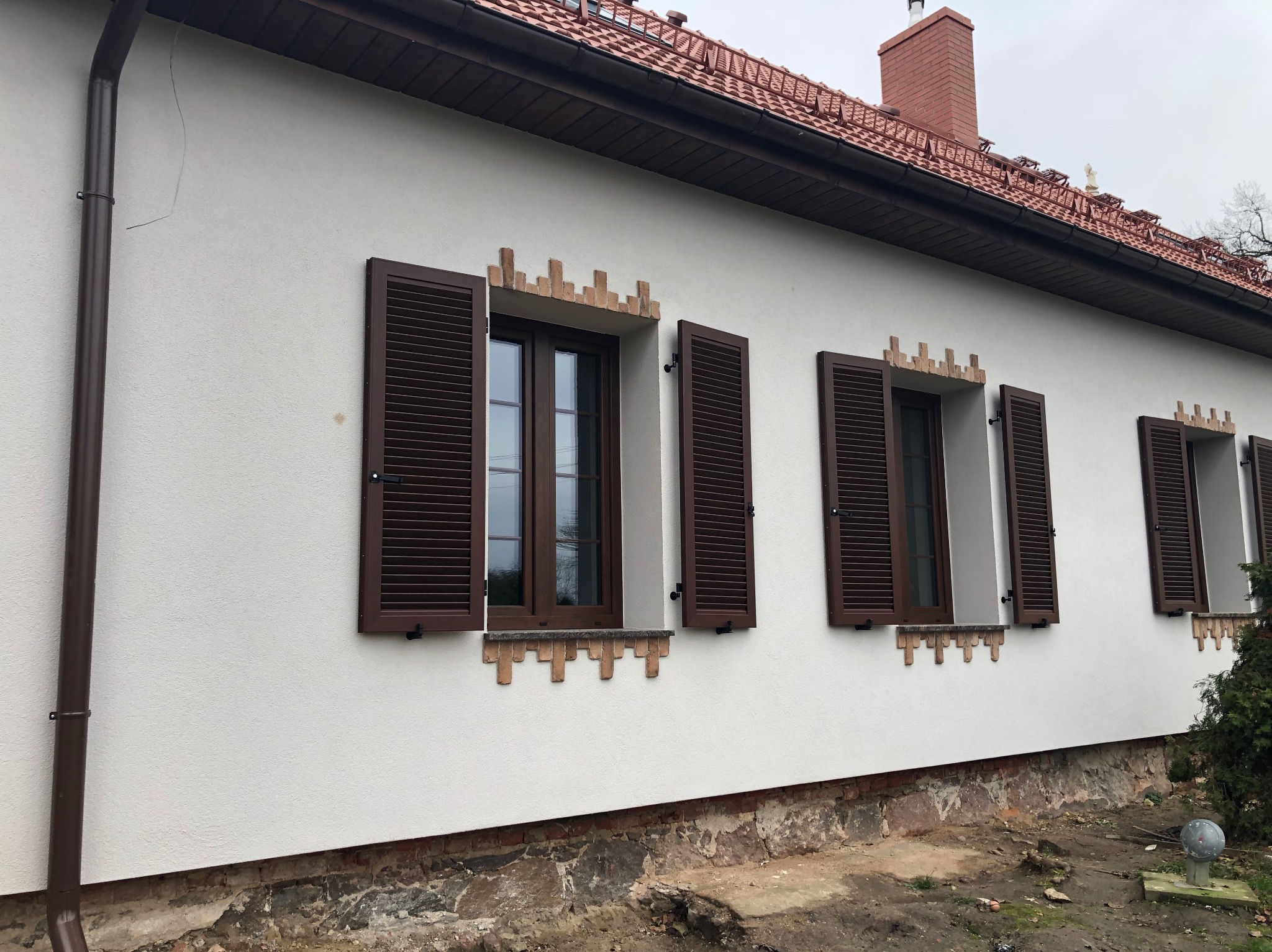 Biała elewacja domu z brązowymi okiennicami i ceglanymi zdobieniami nad oknami. Widoczny fragment dachu z czerwoną dachówką i brązowa rynna.