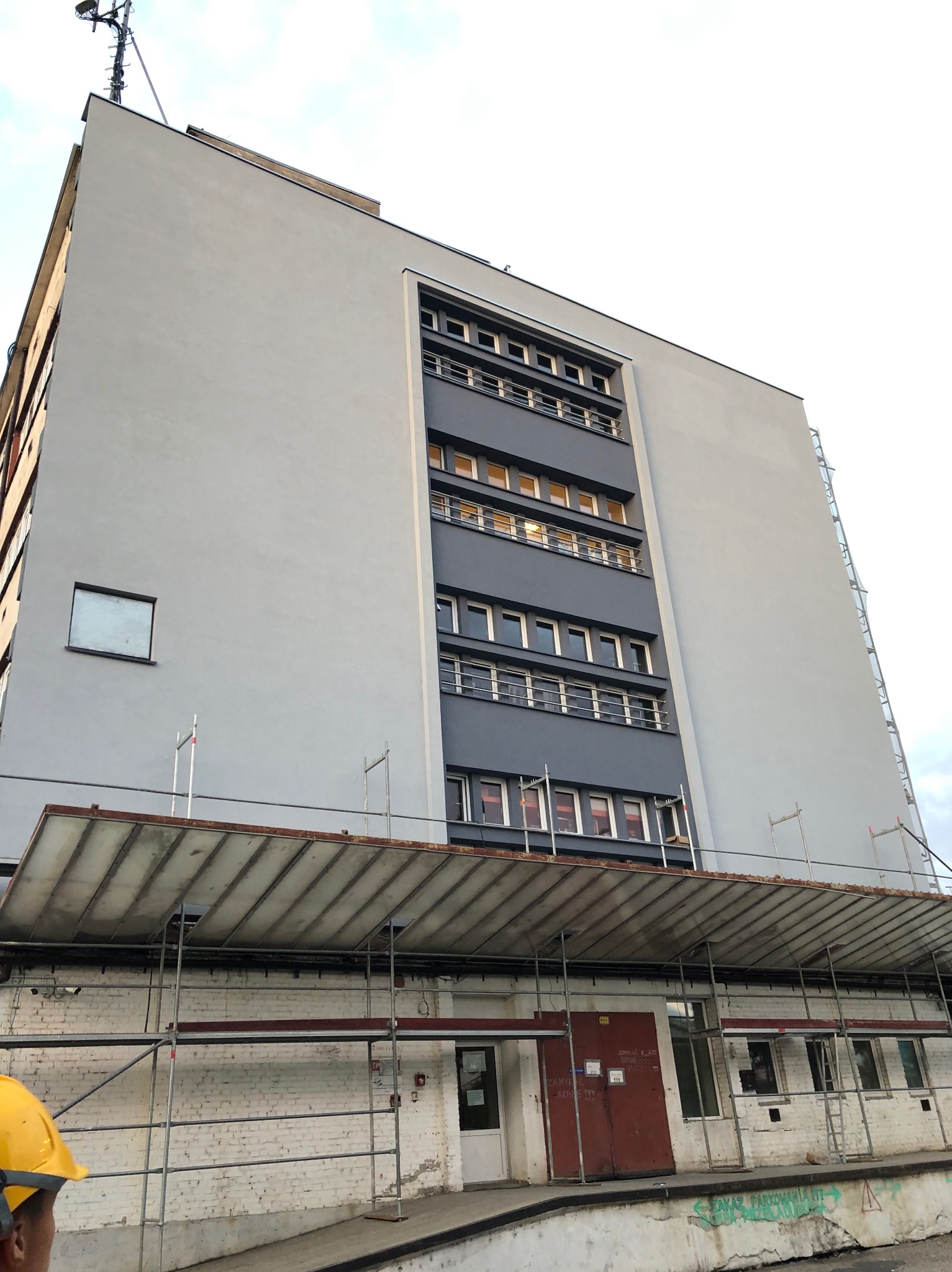 Wysoki budynek po renowacji elewacji, widoczne rusztowania i fragment osoby w kasku. Szara, odnowiona fasada z ciemnoszarym pionowym pasem okiennym.