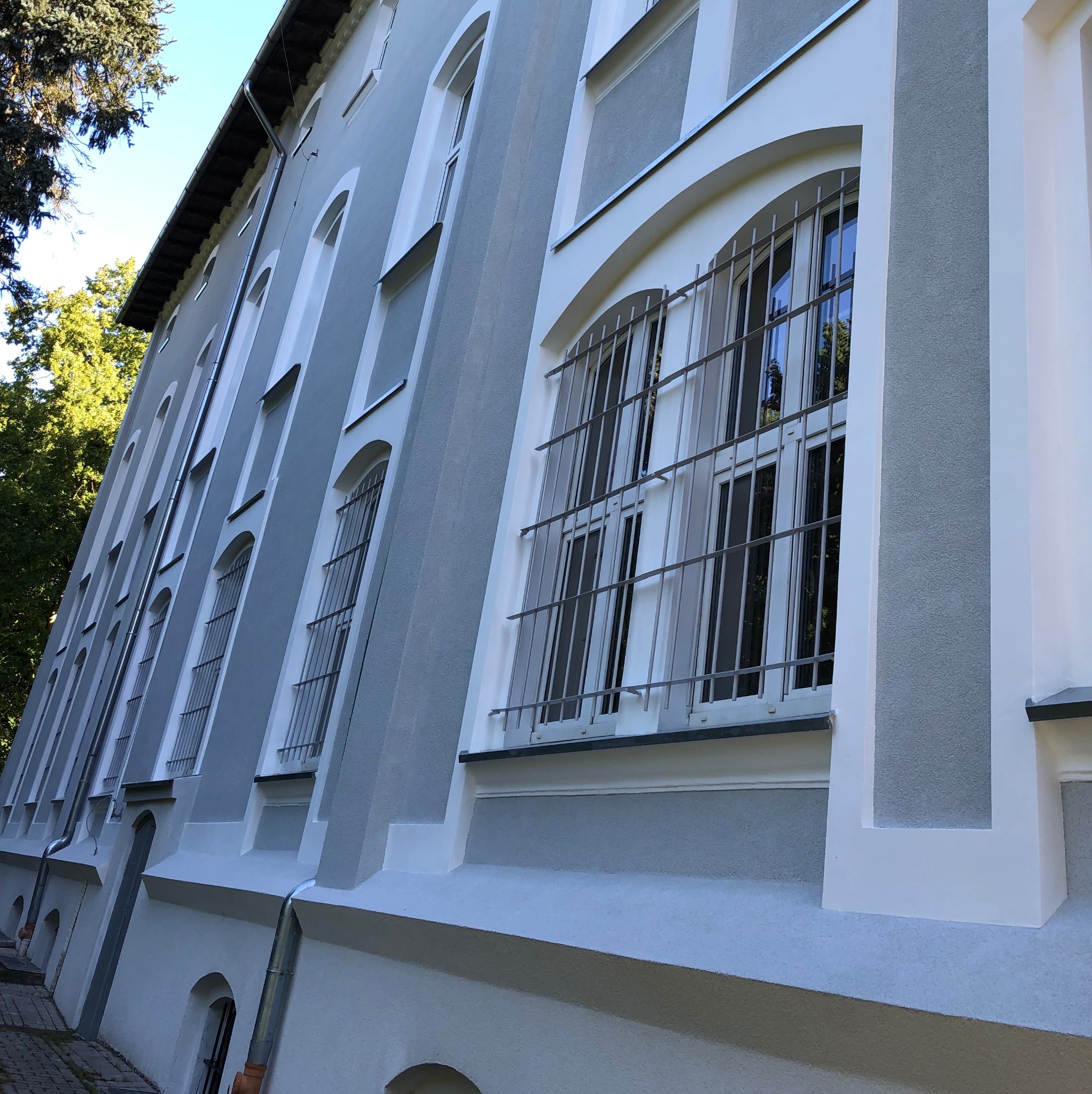Odnowiona elewacja budynku z szarymi ścianami i białymi detalami architektonicznymi. Okna z kratami, widok z dołu, fragment drzewa w tle.