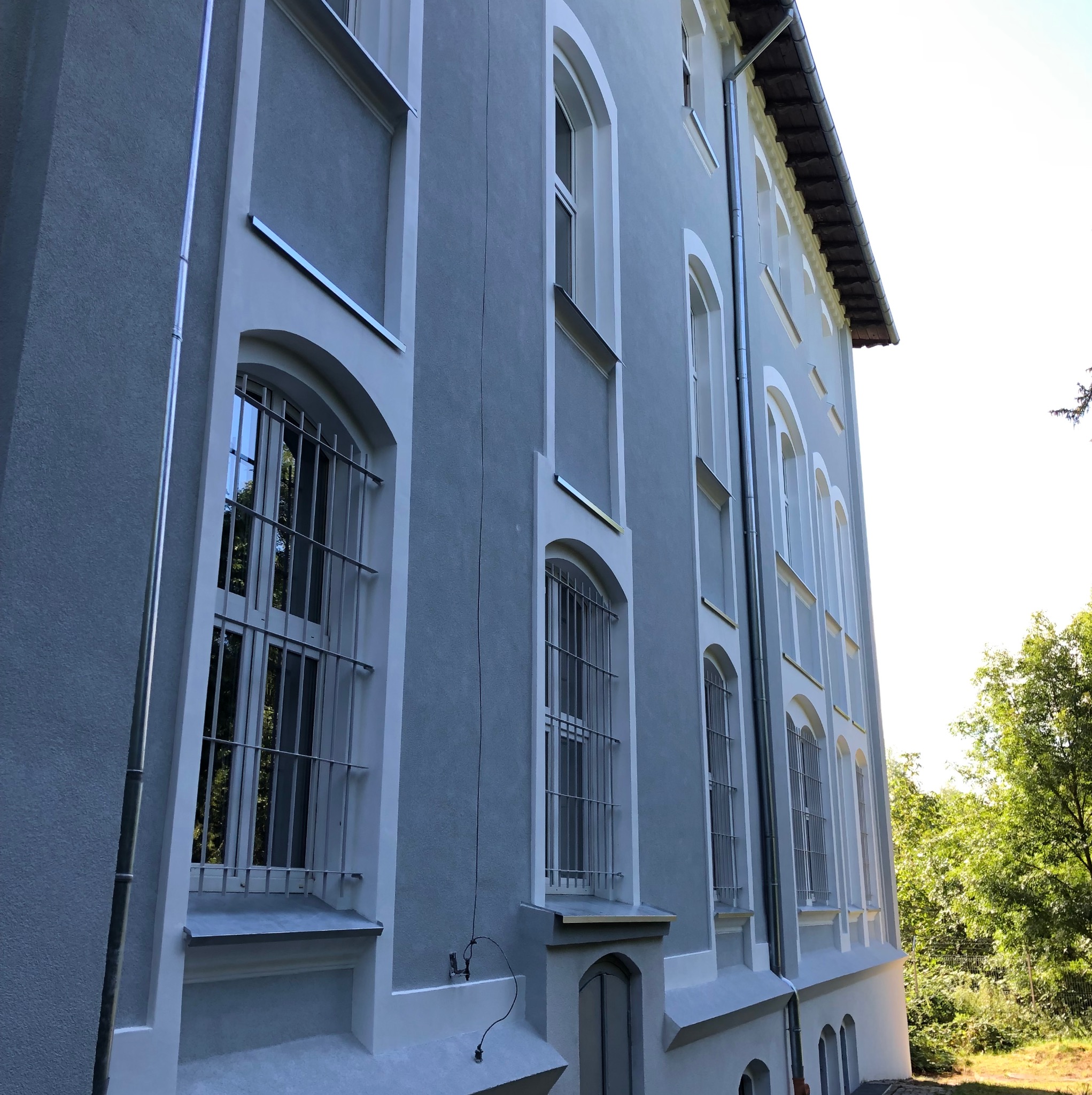 Odnowiona elewacja budynku z szarym tynkiem i białymi detalami architektonicznymi, widok z dołu, z oknami z kratami i rynnami. Świeżo pomalowana fasada.