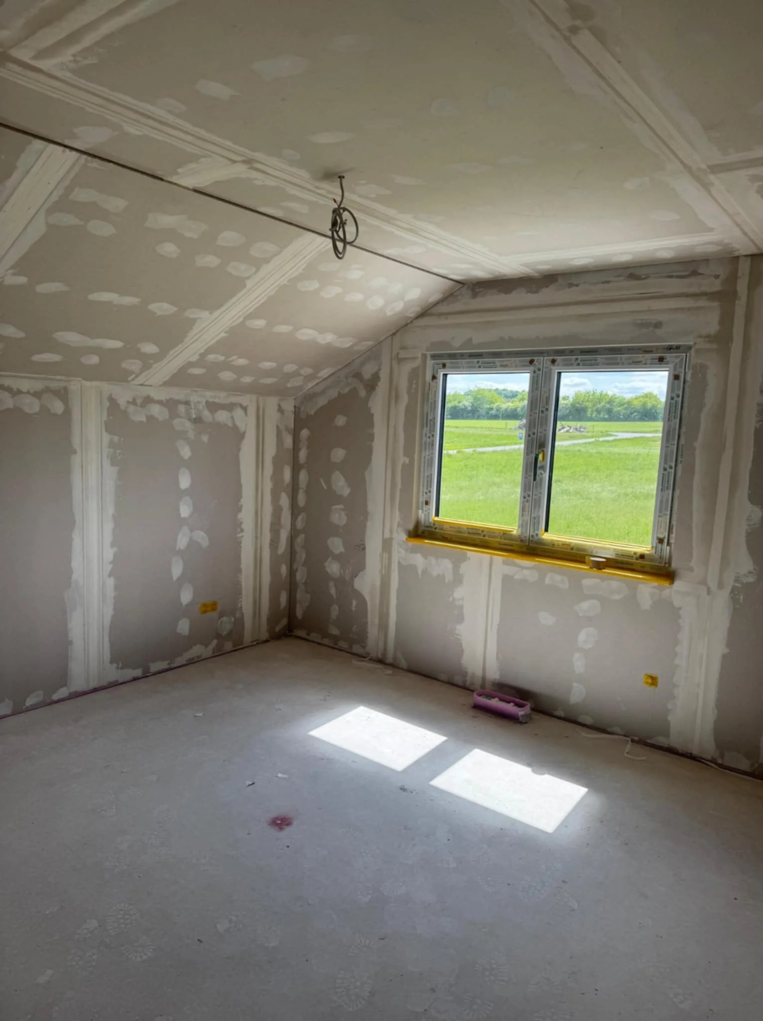 Pokój na poddaszu w trakcie prac wykończeniowych. Ściany i sufit pokryte płytami gipsowo-kartonowymi, widoczne szpachlowanie. Okno z widokiem na zielony krajobraz.